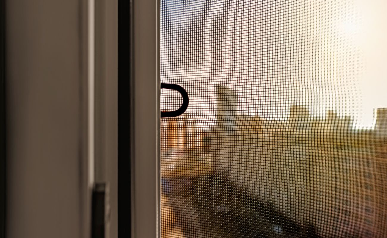 Fenster mit einem Insektenschutzgitter, im Hintergrund sind unscharfe Stadtgebäude und ein heller Himmel zu sehen.